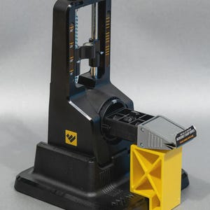 Könnte beinhalten: Schwarzer Messerschärfer mit gelber Halterung und grauem Schärfführer. Der Schärfer hat eine vertikale Skala und einen Knopf oben. Die Basis hat ein Logo. Der Text auf dem Führer lautet "INCREDIBLY SHARP KNIVES".