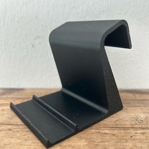 Könnte beinhalten: Ein schwarzer Telefonständer mit strukturierter Oberfläche. Der Ständer hat eine Basis mit einer Nut und eine gebogene Stütze. Der Ständer besteht aus einem soliden Material und ist für ein Mobiltelefon konzipiert.