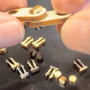May include: A close-up of a wooden craft kit with small wooden pieces shaped like arrows. The pieces are scattered on a black surface. A hand is holding a piece of the kit.