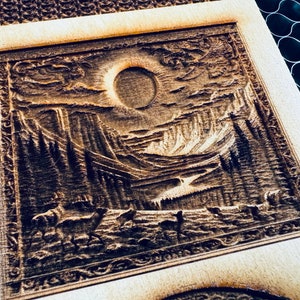 Puede incluir: Una placa de madera con una imagen tallada de un paisaje montañoso con un gran sol en el cielo. El sol está parcialmente oscurecido por nubes. Hay árboles y un río en primer plano.