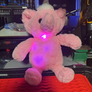 May include: A pink plush elephant toy with a party hat and a glowing light on its chest. The toy is soft and fluffy, with a trunk, ears, and small arms and legs. The hat is silver and white, and the light glows purple.