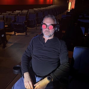 May include: A man wearing a black shirt and blue jeans sits in a theater seat. He is wearing red-tinted goggles and headphones.