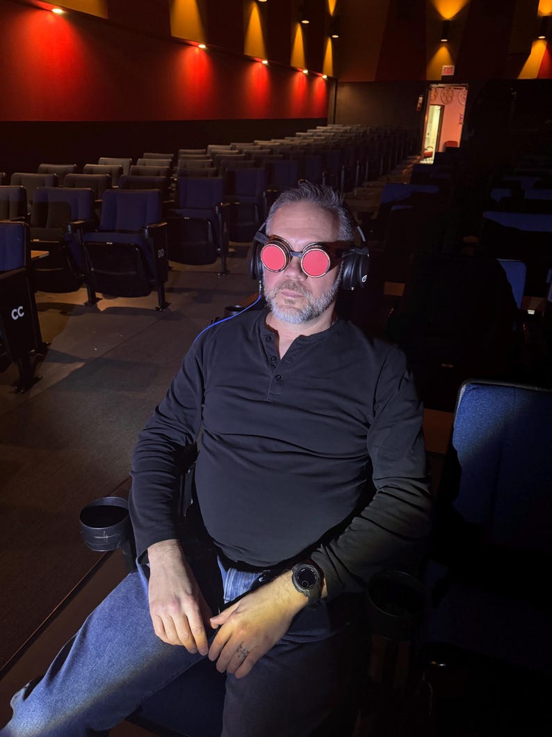 May include: A man wearing a black shirt and blue jeans sits in a theater seat. He is wearing red-tinted goggles and headphones.