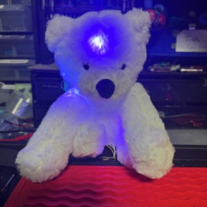 May include: A white plush teddy bear with a glowing blue light emanating from its head. The bear has a black nose and is sitting on a red surface. Wires are visible, suggesting an electronic component.