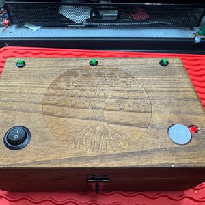 Puede incluir: Una caja de madera con un diseño de árbol de la vida grabado en la tapa. La caja tiene un interruptor de encendido/apagado negro, un botón gris y una luz roja. También se ven tres luces verdes en la parte superior.
