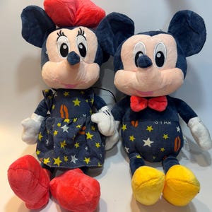 May include: Two plush Disney character dolls. One is Minnie Mouse, wearing a blue dress with yellow stars and a red bow. The other is Mickey Mouse, wearing a blue outfit with yellow stars and a red bow tie. Both have white gloves and large ears.