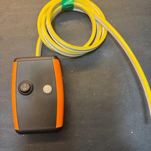 Puede incluir: Un dispositivo electrónico negro y naranja con dos botones y un cable amarillo y gris. El cable está enrollado y asegurado con cinta verde. El dispositivo tiene un botón de encendido/apagado y un botón más pequeño.
