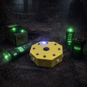 Op de afbeelding: Een verzameling spokenjachtapparatuur, waaronder een geel achthoekig apparaat met een centrale knop, een cilindrische zaklamp met een groen digitaal display, een rechthoekige doos met groene lampjes en twee cilindrische sensoren met groene tekst en REM-labels.