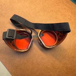 May include: A pair of black leather steampunk goggles with orange lenses and a black adjustable strap.