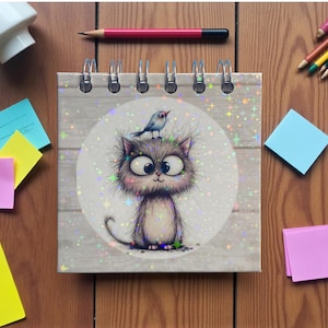 May include: A spiral-bound notebook with a whimsical illustration of a cat with a bird on its head. The cat has large eyes and a fluffy appearance. The notebook is surrounded by colorful sticky notes, pencils, and a pencil.