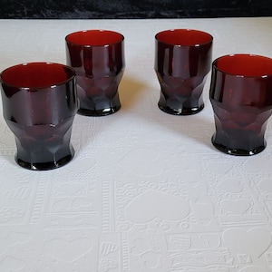 May include: Set of four dark red glass tumblers with a faceted design. The glasses are on a white surface with a textured pattern.