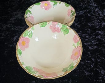 May include: Two off-white ceramic bowls with a floral design. Each bowl features pink flowers with yellow centers and green leaves around the rim. The rims have a gold trim. The bowls are stacked on top of each other.