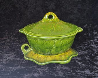 May include: A green ceramic serving dish with a lid. The dish is decorated with a leaf pattern and has a matching base.