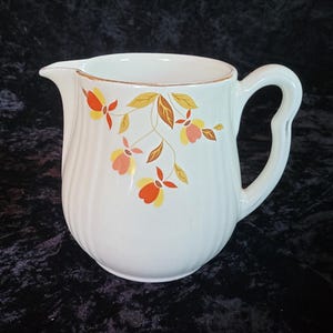May include: White ceramic pitcher with a floral design featuring orange, yellow, and brown leaves. The pitcher has a rounded body and a handle.