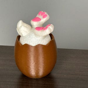 May include: A brown eggshell with a white bunny emerging, showing its fluffy tail and pink-padded feet. The Easter-themed decoration is set on a dark wooden surface, creating a playful and festive scene.