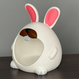 May include: A white, egg-shaped bunny-themed container with pink-lined ears, brown nose, and a light blue tooth. The container has a large opening and small feet. The design is whimsical and playful.