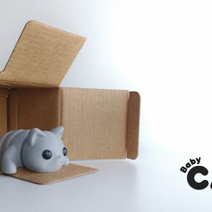 Op de afbeelding: Een grijs, cartoonachtig babykattenfiguurtje bevindt zich in een open kartonnen doos. De kat is opgerold en heeft grote ogen. De tekst "Baby CAT" is in zwart op een witte achtergrond gedrukt.