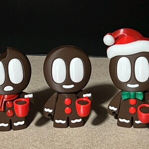 May include: Three gingerbread men figurines, each holding a red mug. One gingerbread man is wearing a red and white Santa hat. The gingerbread men are brown with white eyes and smiles.