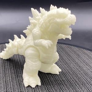 Op de afbeelding: Een wit, in het donker lichtend Godzilla-figuur van plastic. Het figuur staat met open mond en opgeheven armen.