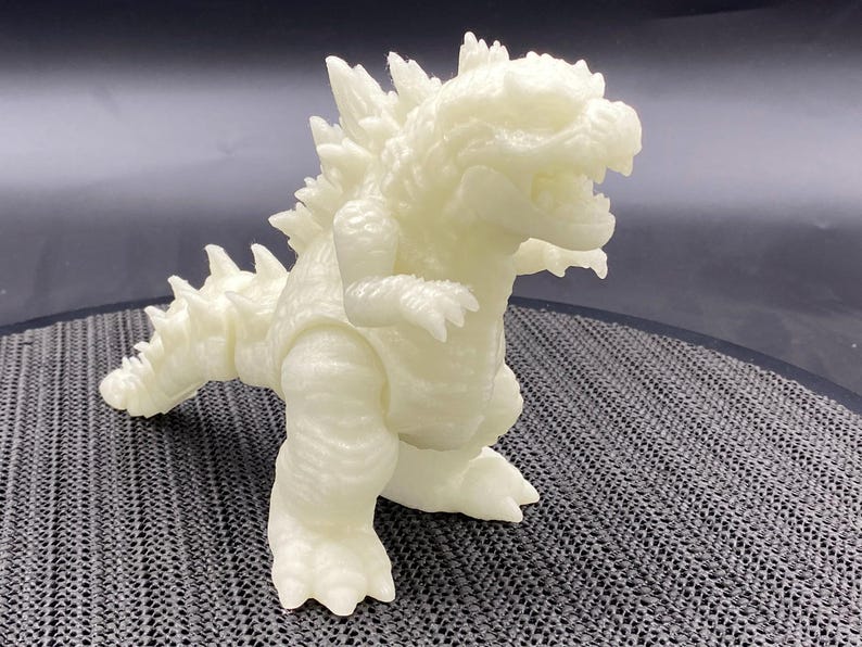 Op de afbeelding: Een wit, in het donker lichtend Godzilla-figuur van plastic. Het figuur staat met open mond en opgeheven armen.