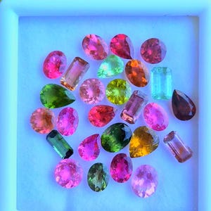 Könnte beinhalten: Eine Sammlung farbiger Edelsteine in einer weißen quadratischen Schale. Die Edelsteine haben verschiedene Formen und Größen, darunter runde, tropfenförmige und rechteckige Schnitte. Zu den Farben gehören Rosa, Grün, Gelb und Braun. Die Schale wird in einer Hand gehalten.