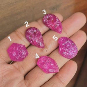 Puede incluir: Cinco gemas talladas de color fucsia con diseños florales y geométricos. Las gemas tienen varias formas, incluyendo lágrima y hexagonal, y están numeradas del 1 al 5. Las gemas se muestran sobre una superficie marrón claro.
