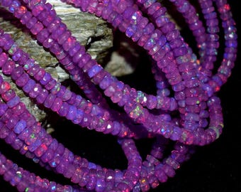 Peut inclure: Plusieurs rangées de perles d'opale violettes facettées. Les perles présentent une iridescence subtile, avec des éclats de vert, de rouge et de bleu. Les perles sont enfilées ensemble, créant un aspect fluide et texturé. Les perles reposent sur une surface sombre.