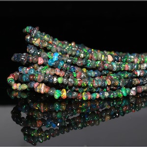 Puede incluir: Hebras de cuentas de piedras preciosas de ópalo negro. Las cuentas son una mezcla de colores, incluyendo verde, rojo, azul y amarillo. Las cuentas son pequeñas e irregulares en forma, y están ensartadas juntas. Las cuentas se muestran sobre un fondo negro.