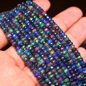 May include: Strands of faceted black opal beads with vibrant flashes of color, including blue, green, and purple. The beads are held in a hand, showcasing their polished surfaces and intricate cuts. These beads are perfect for jewelry making.