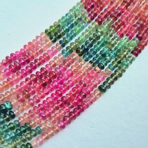 Puede incluir: Cadenas de cuentas de turmalina coloridas en tonos rosa, rojo, verde y azul. Las cuentas son pequeñas y redondas, ensartadas para crear una vibrante exhibición de piedras preciosas naturales. Las cuentas están dispuestas diagonalmente sobre una superficie blanca.