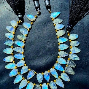 May include: Two strands of gemstone necklaces with leaf-shaped pendants. The pendants have a blue and white color gradient. The necklaces are strung with gold-colored beads and have black tassel accents.