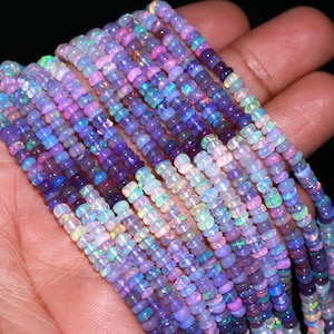 Könnte beinhalten: Stränge schillernder Opalperlen in Lila-, Blau- und Weißtönen. Die Perlen sind klein und rund, mit einem dezenten Schimmer. Die Perlen werden in einer Hand gehalten, um ihre Farbvariationen zu zeigen.