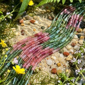 May include: A collection of multi-strand beads featuring cylindrical shapes in varying shades of translucent green and pink. The beads are arranged in parallel lines, showcasing a gradient of colour transitions from deep forest green to soft rose.