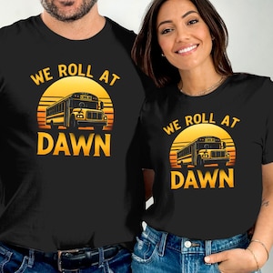 Könnte beinhalten: Ein schwarzes Kurzarm-T-Shirt mit einem Grafikdruck eines gelben Schulbusses vor einem Sonnenuntergang. Der Text "WE ROLL AT DAWN" ist in gelben Großbuchstaben über und unter der Busillustration auf der Brust aufgedruckt.