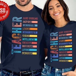 Op de afbeelding: Twee marineblauwe T-shirts met het woord "TEACHER" verticaal in regenboogkleuren gedrukt. Het woord wordt omringd door een lijst met positieve adjectieven die een leraar beschrijven, zoals "hardwerkend", "geïnspireerd", "memorabel", "leider", "resourcevol", "zorgzaam", "intelligent", "creatief", "gemotiveerd", "liefhebbend" en "toegewijd".