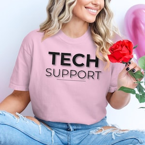 Op de afbeelding: Een roze T-shirt met de tekst "TECH SUPPORT" in het zwart op de voorkant. Het shirt wordt gedragen door een persoon met lang blond haar.