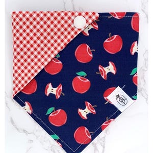 May include: A reversible dog bandana with a red and white gingham pattern on one side and a navy blue side with red apple illustrations. The bandana is secured with a white button and features a small tag.