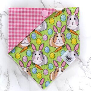 Pode incluir: Bandana reversível para animais de estimação. Um lado apresenta um padrão xadrez rosa, o outro um design primaveril. O lado primaveril apresenta porquinhos-da-índia com orelhas de coelho, ovos de Páscoa e cenouras sobre um fundo verde.