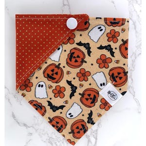 Pode incluir: Bandana para cães com tema de Halloween, com fundo bege com abóboras laranja, flores, fantasmas e morcegos. O canto superior esquerdo é laranja sólido com bolinhas brancas. A bandana é presa com um botão branco.