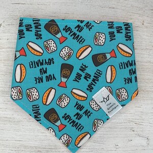 May include: A turquoise pet bandana with a repeating pattern of sushi, soy sauce bottles, and the text "YOU ARE MY SOYMATE!". The bandana has a triangular shape and a small white tag with a crown logo and text.