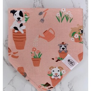 May include: Pink bandana with a pattern of dogs in flower pots, watering cans, and gardening tools. The bandana has a white label with a pineapple logo.
