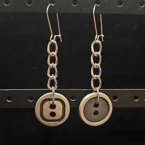 May include: A pair of silver earrings with a button charm. Each button has a different design, one with a smooth surface and the other with a ribbed surface. The buttons are attached to a silver chain.