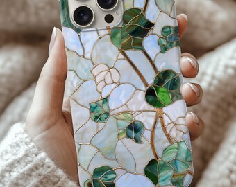 May include: A decorative phone case featuring a stained glass design with green leaves and white flowers. The case has a gold outline and a multi-camera lens cutout. The background is a soft, neutral color.