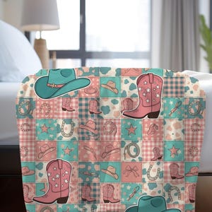 May include: A patchwork throw blanket featuring a repeating pattern of cowboy hats, boots, horseshoes, and stars in shades of pink, teal, and white. The blanket is draped over a bed.