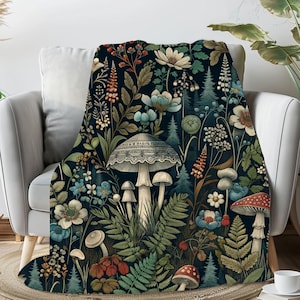 Puede incluir: Una manta de tiro con un fondo oscuro que presenta un patrón repetitivo de setas coloridas, flores y helechos. Las setas son blancas con manchas marrones y tienen un diseño de encaje en la parte superior.