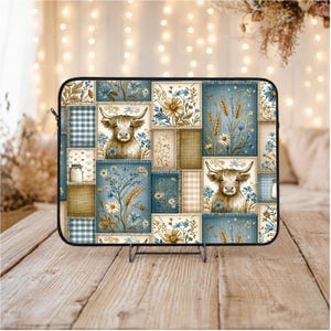 Op de afbeelding: Een laptoptas met een patchwork-ontwerp met een Schotse hooglander, bloemen en tarwearen. De hoes heeft een zwarte rand en wordt gepresenteerd op een houten oppervlak. Het kleurenpalet omvat blauw, bruin en crème.