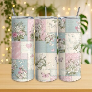 Op de afbeelding: Drie geïsoleerde bekers met deksels en rietjes. Elk heeft een patchwork ontwerp met bloemboeketten, vlinders en harten in pastelroze, blauwe en crèmekleuren. Metrische maten.