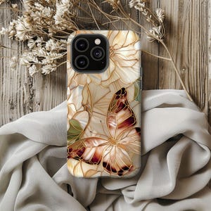 May include: A phone case featuring a butterfly and floral design. The case has a cream-colored background with a stained-glass effect. The butterfly has brown and gold details. The case is on a wooden surface with dried flowers and a draped cloth.