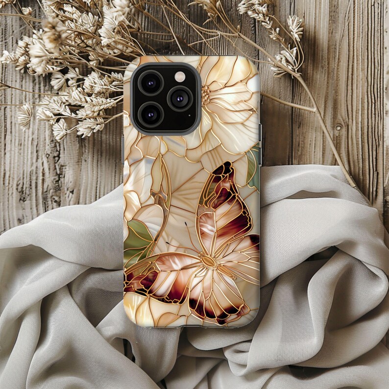 May include: A phone case featuring a butterfly and floral design. The case has a cream-colored background with a stained-glass effect. The butterfly has brown and gold details. The case is on a wooden surface with dried flowers and a draped cloth.