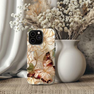 May include: A phone case featuring a butterfly and floral design. The case has a cream-colored background with gold outlines and accents. The butterfly has brown and cream-colored wings. The case is next to a white vase with dried flowers.